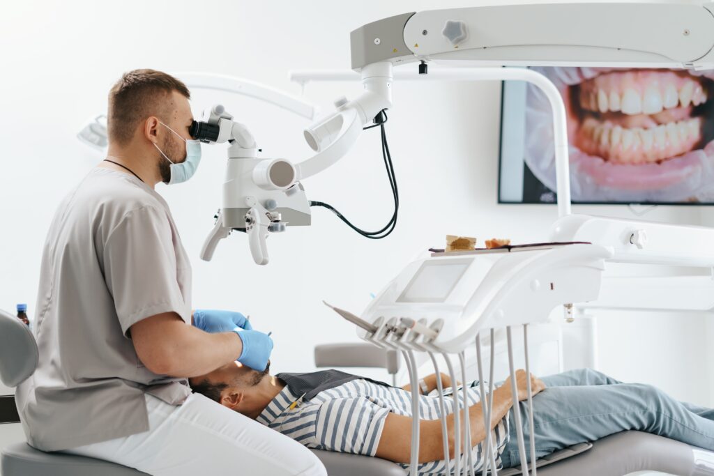 young-man-with-patient-bib-dental-chair-dentist-who-sits-him-he-looks-his-teeth-using-dental-microscope-holds-dental-bur-mirror-min