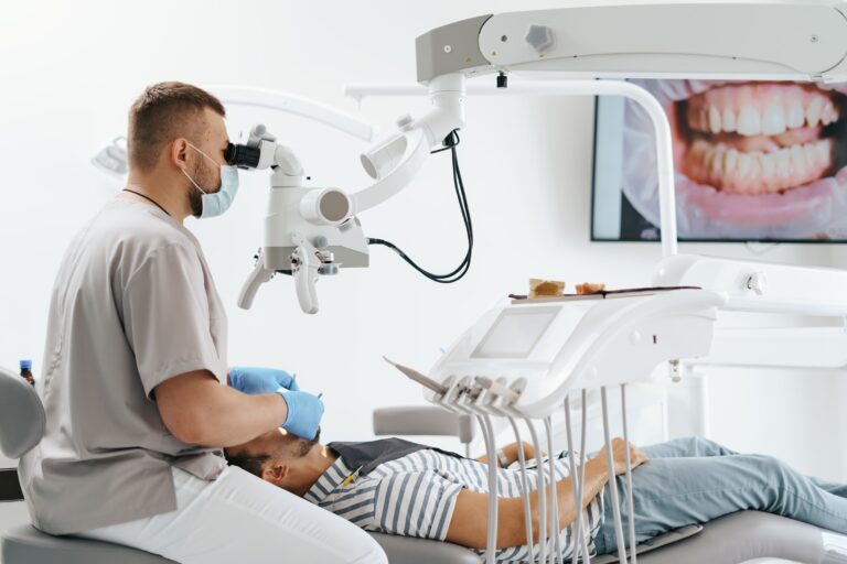 young-man-with-patient-bib-dental-chair-dentist-who-sits-him-he-looks-his-teeth-using-dental-microscope-holds-dental-bur-mirror-min